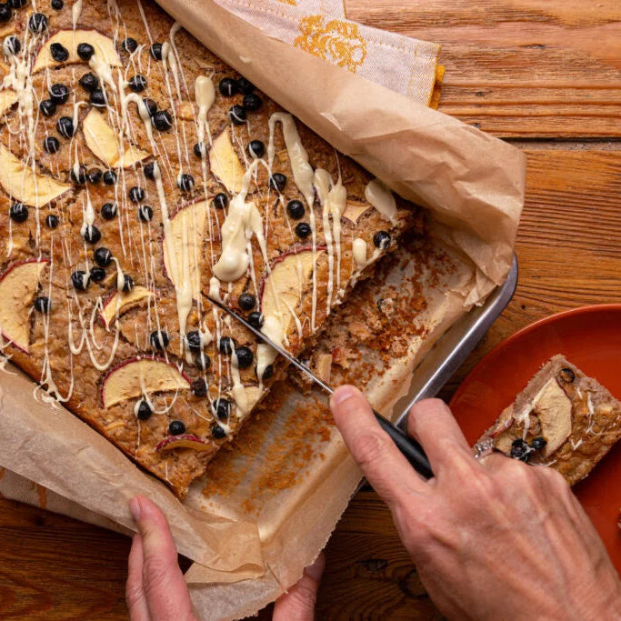 Apfelkuchen mit Schwarze Johannisbeeren und Zimt auf einem Holztisch, wobei gerade ein Stück herausgenommen wird.