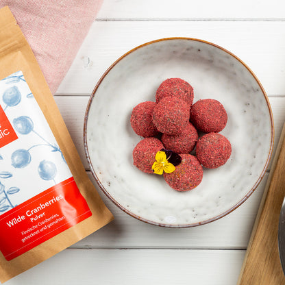 Rote Cranberry-Power-Bälle auf einem weissen Teller mit einer Verpackung von Cranberry-Pulver auf einer Holzoberfläche.
