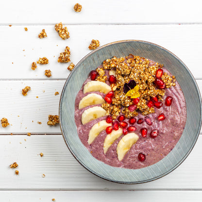 Schüssel mit pinkem Preiselbeeren-Smoothie, garniert mit Sanddorn-Granola, Bananen und Granatapfelkernen auf einer weissen Holzoberfläche.