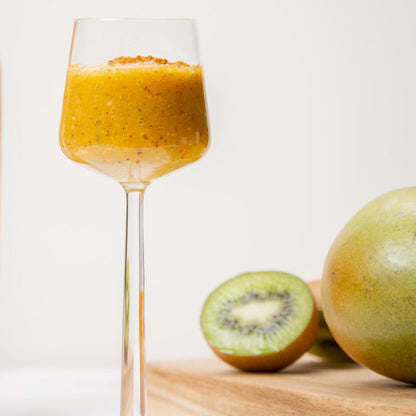 Ein hohes Glas, gefüllt mit einem gelben Hagebutten-Orangen-Getränk, mit einer Kiwi und einer Mango daneben.