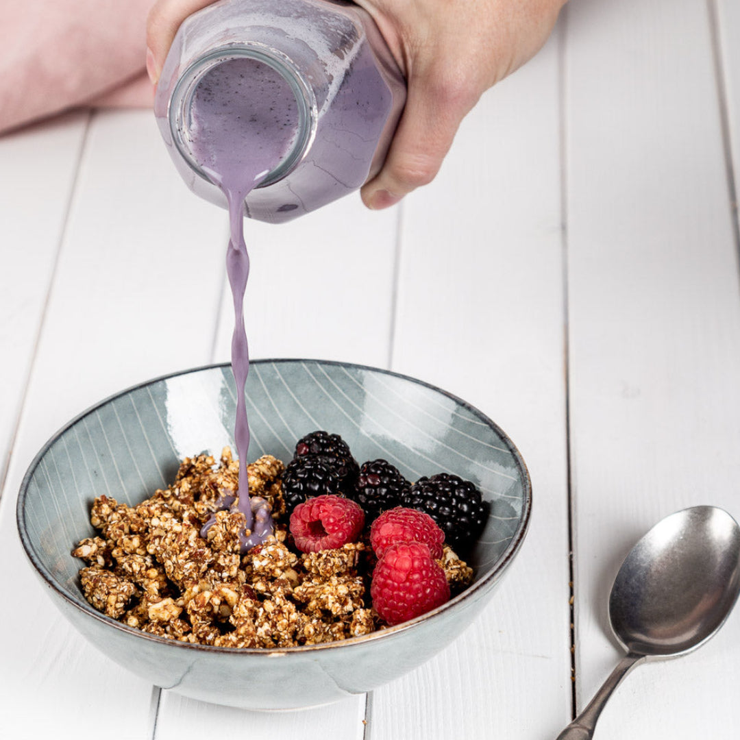 Eine Hand gießt Heidelbeermilch in eine Schüssel mit Sanddorn-Granola und Beeren auf einem weißen Tisch, neben einem silbernen Löffel.
