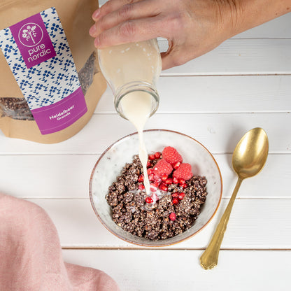 Eine Person gießte ein Milchgetränk aus einer Flasche in eine Schüssel mit Heidelbeer-Granola und Himbeeren, neben einer pure nordic Granola Packung