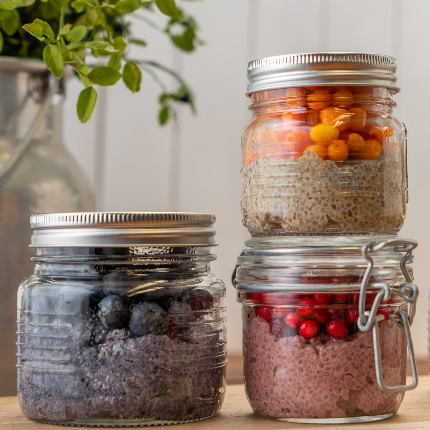 Drei gestapelte Glasbehälter mit Overnight Oats, Chiasamen und Beeren auf einem Tisch, mit einigen Heidelbeerblättern im Hintergrund.