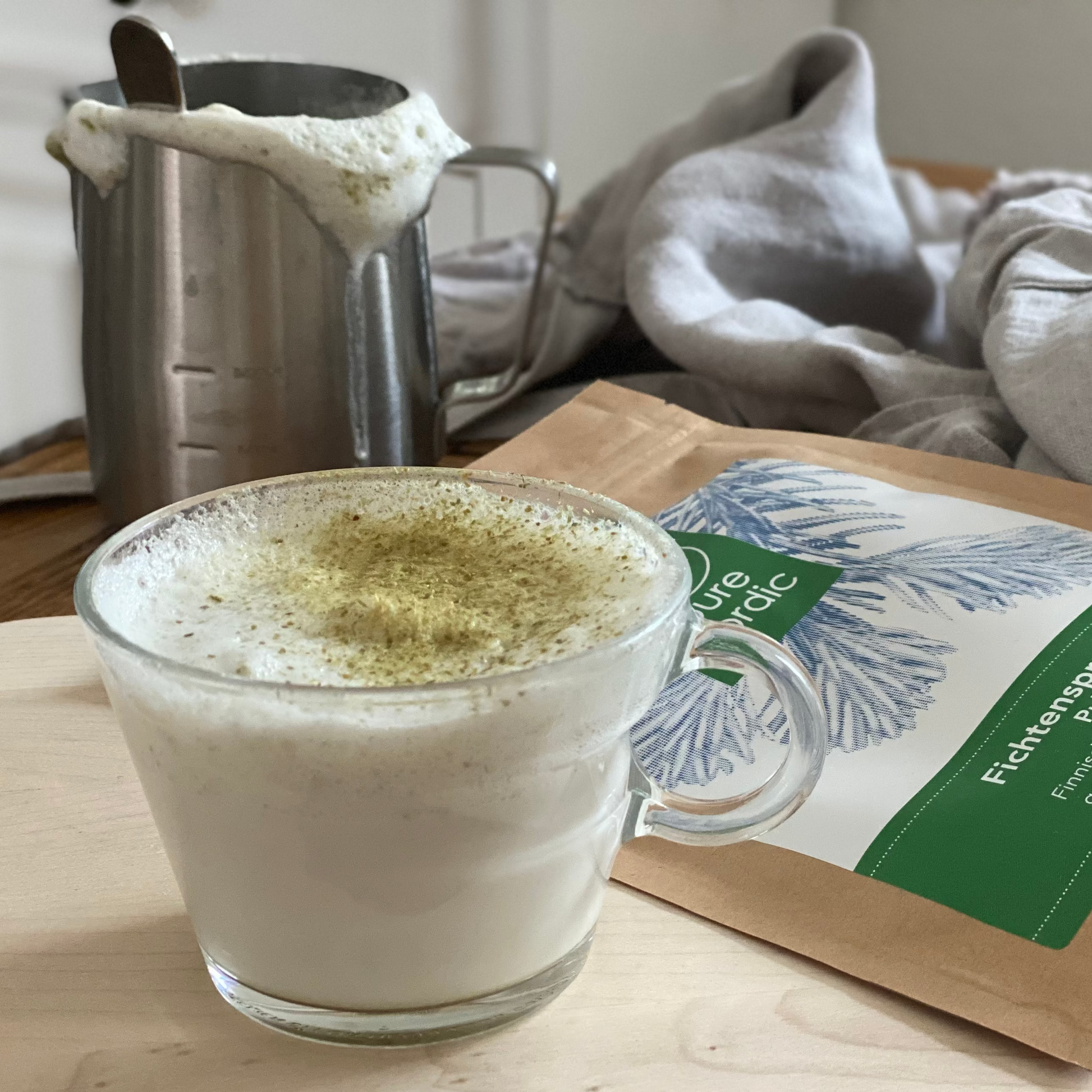 Ein Glas Fichtensprossen-Latte auf einem Tisch, zusammen mit einer Fichtensprossenpulver Packung und einer Kanne mit schäumender Milch.