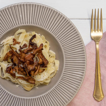Ein weisser Teller Pasta mit Trompetenpfifferlingen auf einem weissen Teller, daneben eine goldene Gabel auf einem Holztisch.