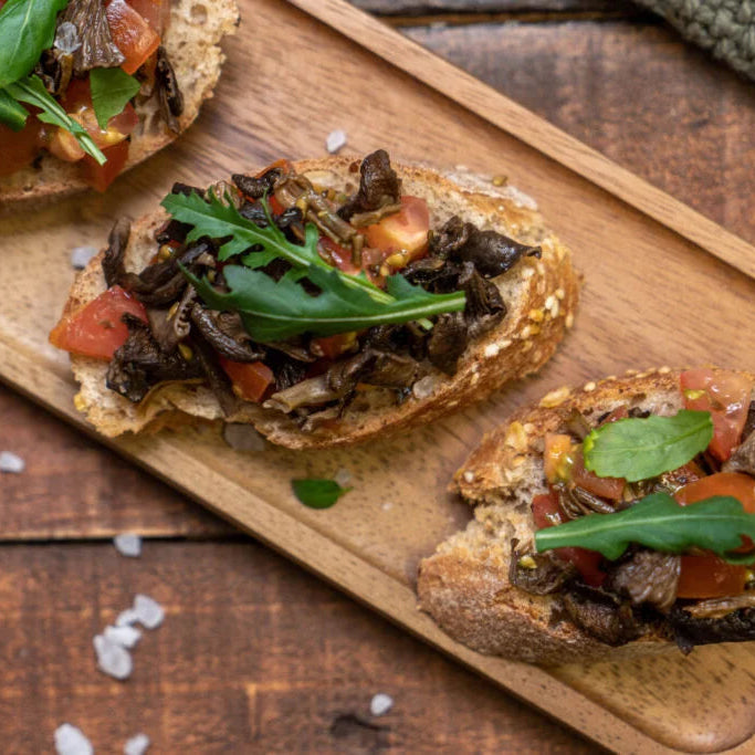 Bruschetta mit Trompetenpfifferlingen, Tomaten und Basilikumblättern auf einem Holzbrett.