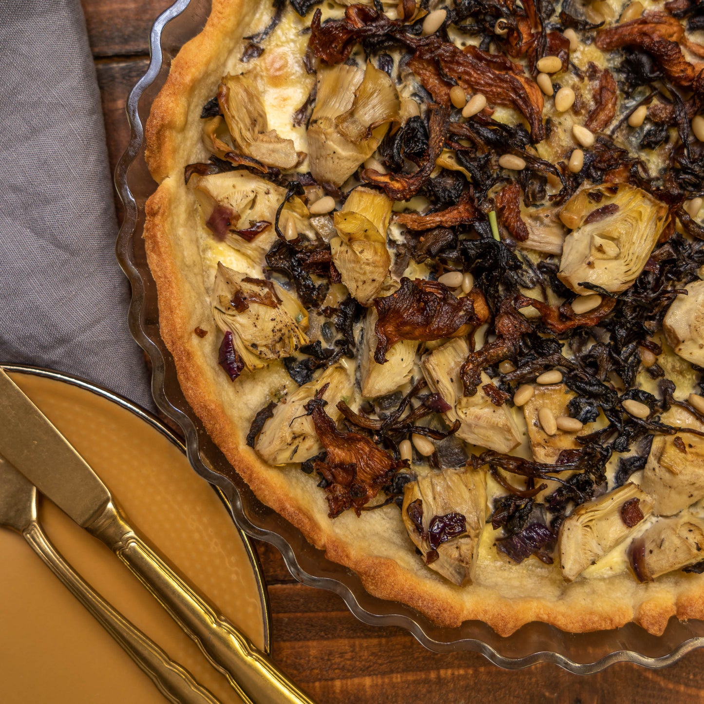 Quiche mit Artischocken, sonnengetrockneten Tomaten und finnischen Pfifferlingen auf einem Holztisch, mit einem gelben Teller sowie einer goldenen Gabel und einem goldenen Messer.