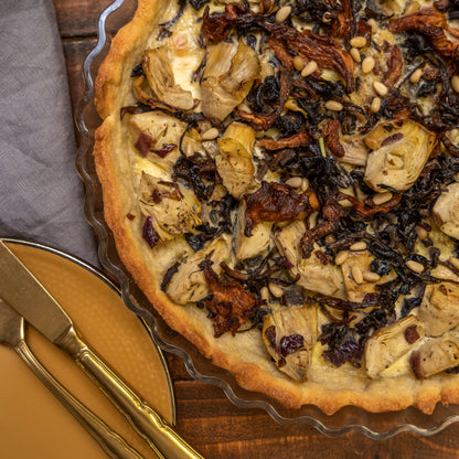 Quiche mit Artischocken, sonnengetrockneten Tomaten und finnischen Pfifferlingen auf einem Holztisch, mit einem gelben Teller sowie einer goldenen Gabel und einem goldenen Messer.