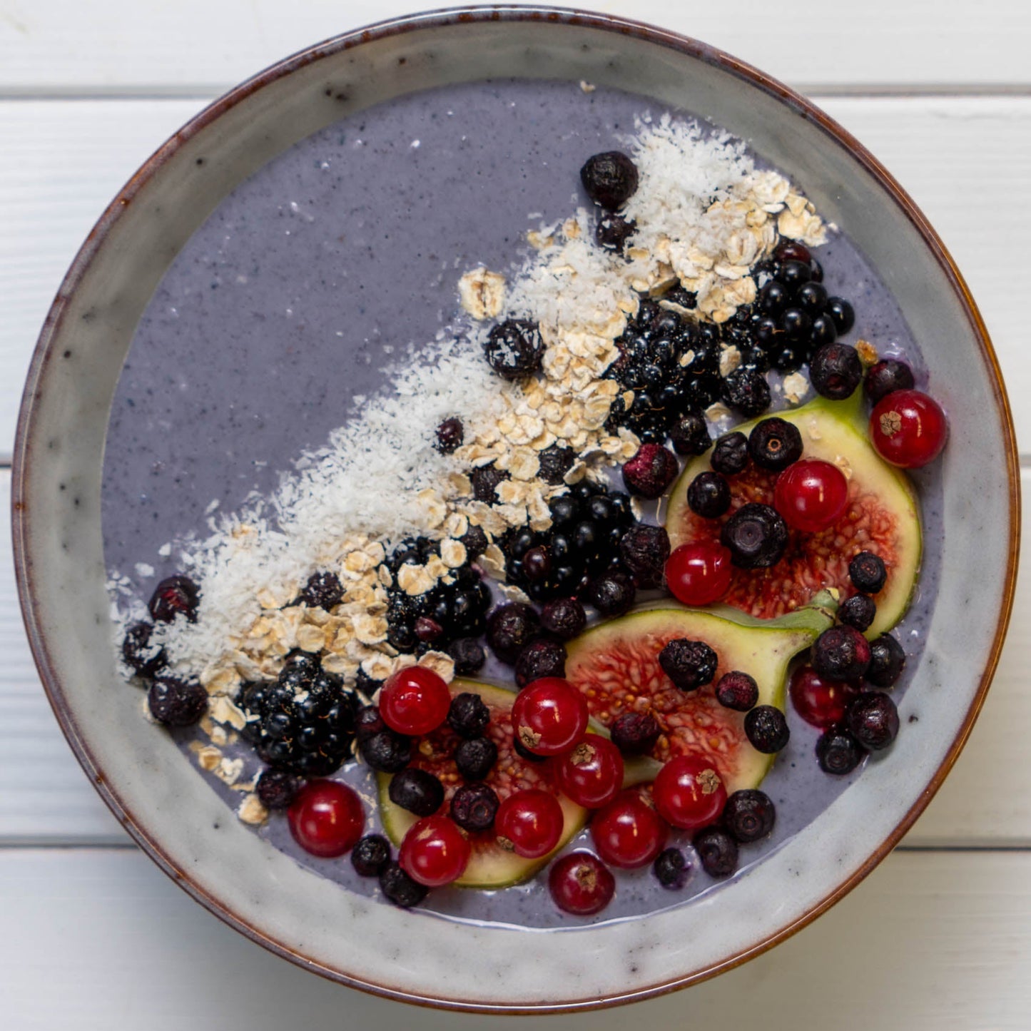 Lila Smoothie-Bowl mit Beeren, Früchten und Haferflocken auf einem weißen Holztisch.