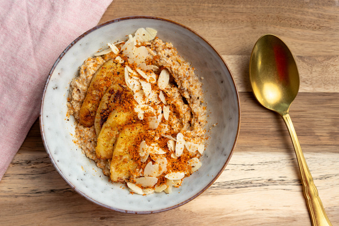 Schüssel mit Porridge, karamellisierten Bananenscheiben, Beerenpulver und Mandeln, daneben ein goldener Löffel.