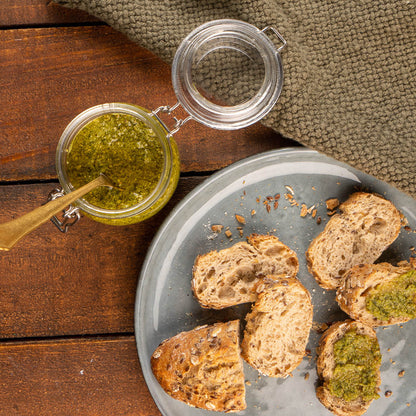 Ein Glas mit grünem Brennnesselpesto mit einem Löffel auf einem Holztisch, neben einem Teller mit Brotscheiben, die mit dem Pesto bestrichen sind.