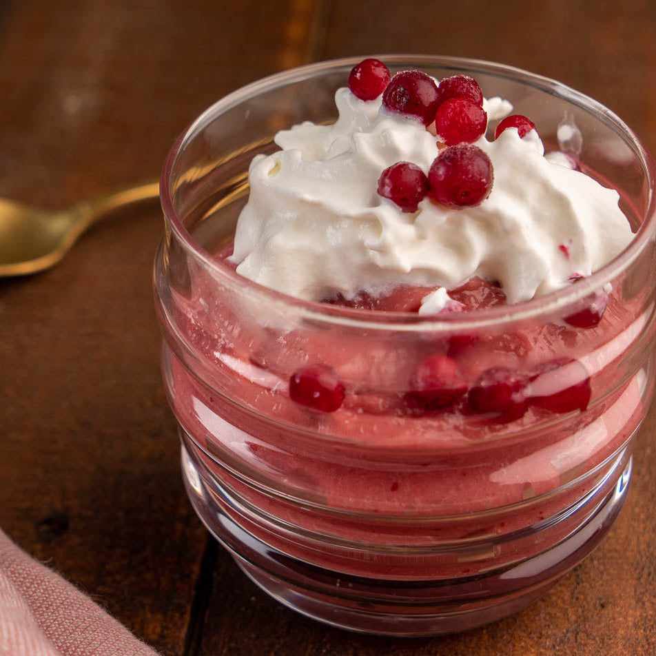 Ein Glas-Dessertbecher mit einem schaumigen Cranberry-Dessert, Schlagsahne und Preiselbeeren auf einem Holztisch mit einem goldenen Löffel.