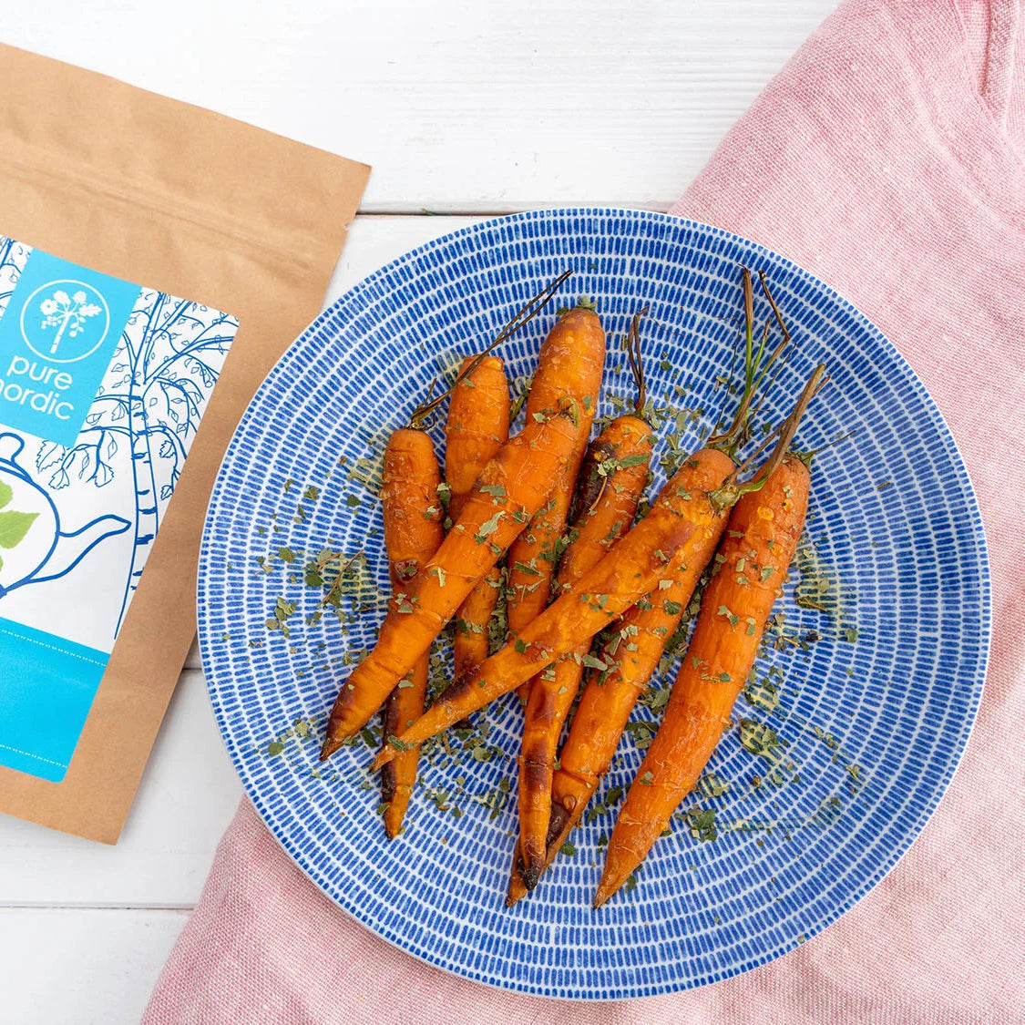 Im Ofen glasierten Karotten, bestreut mit zerkleinerten getrocknete Birkenblättern, auf einem blauen Teller und einer pure nordic Birkenblätter Packung.
