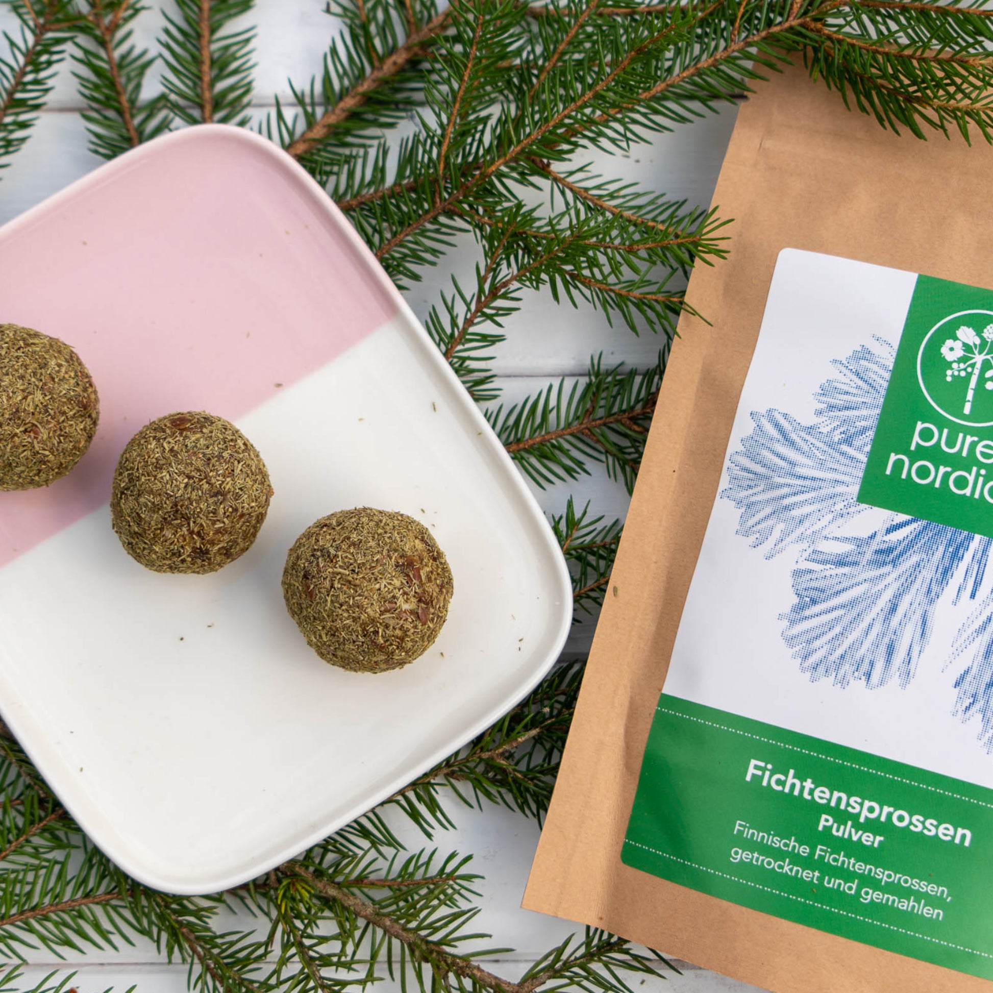 Drei Fichtensprossen-Energiebällchen auf einem weißen Teller mit einer pure nordic Verpackung,  auf einer weißen Holzoberfläche, dekoriert mit Kieferästen.